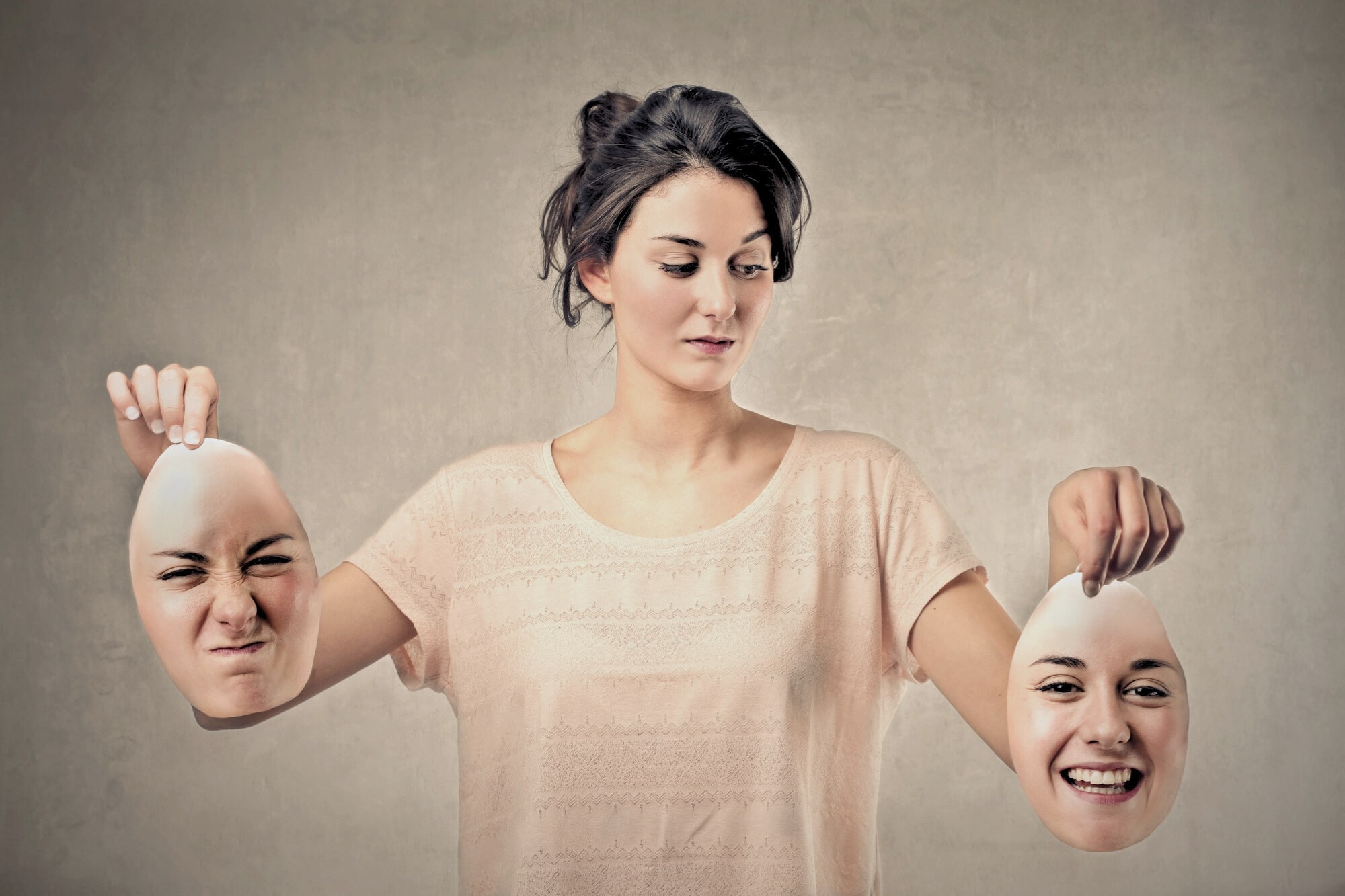 A woman looks down, holding two removable face masks in each hand—one with a scrunched, angry expression, the other with a big, happy smile. She stands against a plain, neutral background.