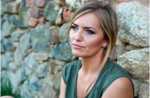 A woman with blonde hair and a green top sits against a stone wall outdoors, looking thoughtfully into the distance with a neutral expression.