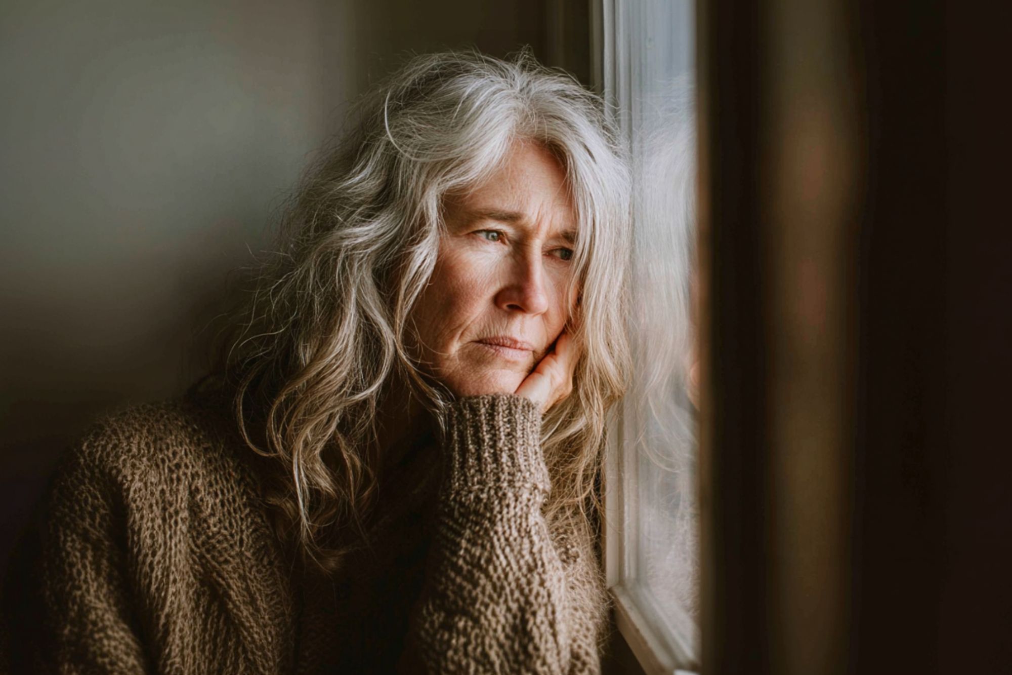An older woman with long gray hair and a brown sweater rests her chin on her hand, gazing thoughtfully out of a window with a pensive expression. Soft natural light illuminates her face.