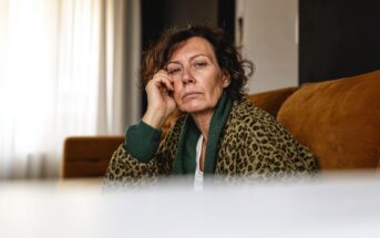 A middle-aged woman with curly hair sits on a brown sofa, resting her face on her hand and looking thoughtfully at the camera. She is wearing a leopard print robe and a green sweater. Light streams in from a window behind her.