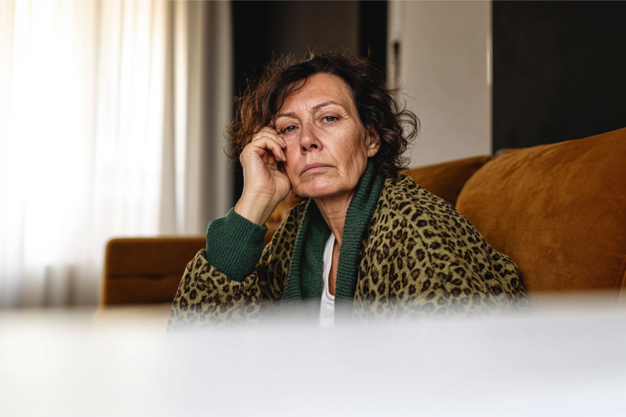 A middle-aged woman with curly hair sits on a brown sofa, resting her face on her hand and looking thoughtfully at the camera. She is wearing a leopard print robe and a green sweater. Light streams in from a window behind her.
