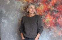 An older woman with gray hair and a gray knit sweater stands in front of a colorful, abstract painted background, looking to the side with a relaxed and thoughtful expression, hands in her pockets.