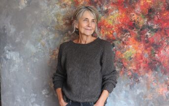 An older woman with gray hair and a gray knit sweater stands in front of a colorful, abstract painted background, looking to the side with a relaxed and thoughtful expression, hands in her pockets.