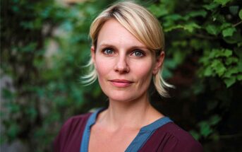 A woman with short blonde hair and green eyes stands outdoors in front of leafy green foliage, wearing a burgundy top with a blue neckline, looking directly at the camera with a calm expression.
