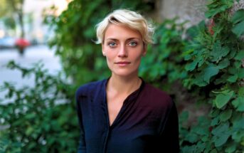 A person with short blonde hair and blue eyes stands outside, wearing a dark shirt. Green leaves and plants are in the background, giving a natural and serene atmosphere. The person looks calmly at the camera.