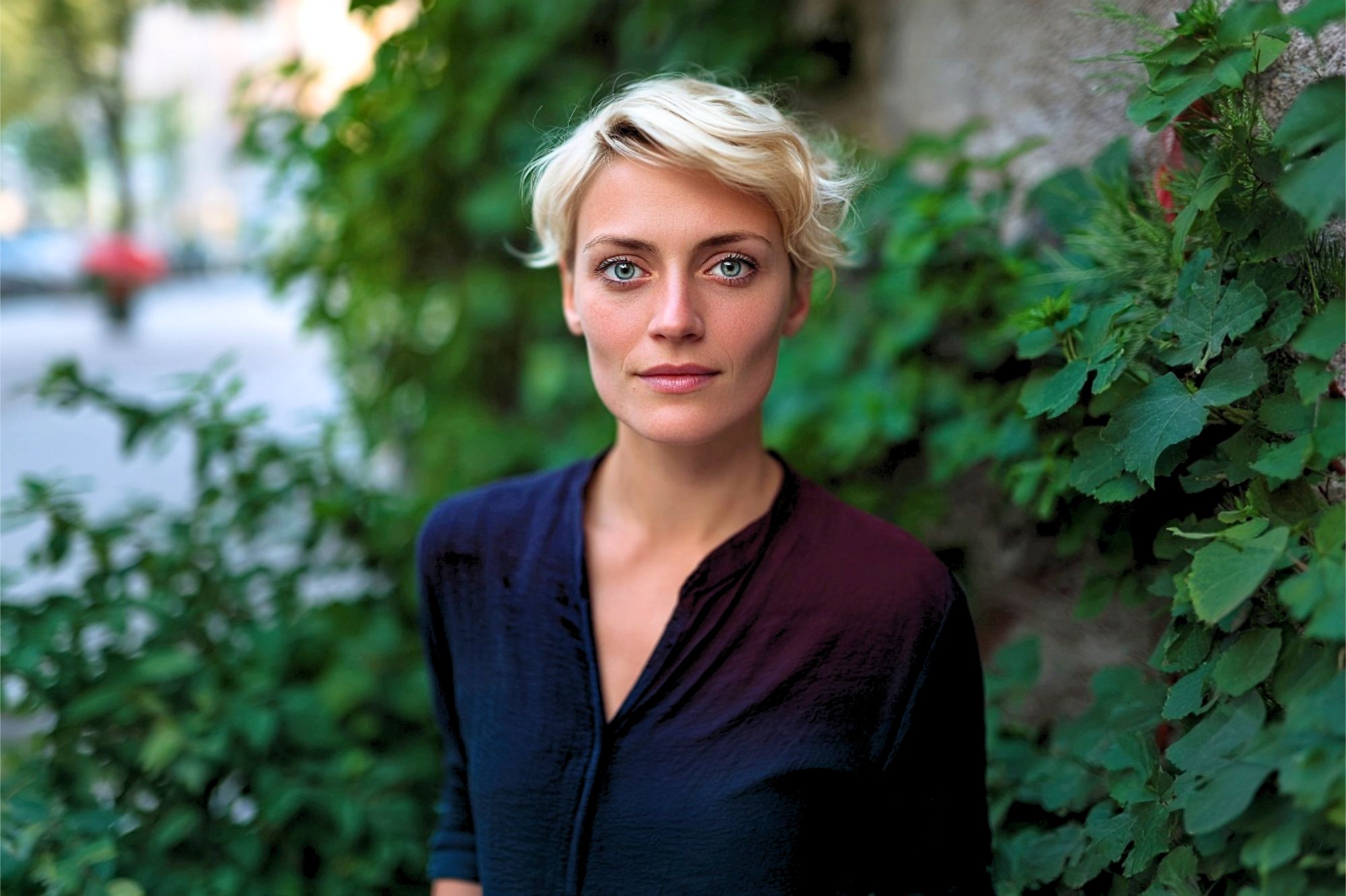A person with short blonde hair and blue eyes stands outside, wearing a dark shirt. Green leaves and plants are in the background, giving a natural and serene atmosphere. The person looks calmly at the camera.