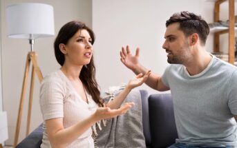 A woman and a man sit on a couch indoors, facing each other and appearing to have an intense argument. Both have raised hands and frustrated facial expressions. A lamp and shelving are visible in the background.