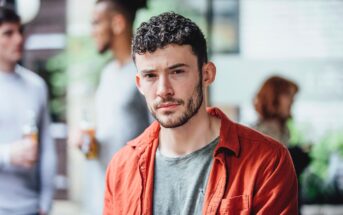 A man with short curly hair and a trimmed beard, wearing a red shirt over a gray t-shirt, looks at the camera. Several people are blurred in the background, standing and talking outdoors.
