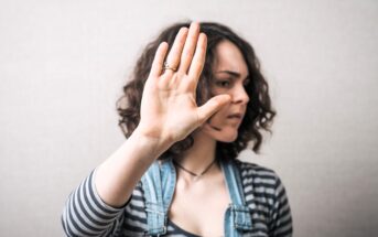 A woman with curly hair wearing a striped shirt and denim overalls holds up her hand in front of her face as if signaling "stop" or "no," with a serious expression and looking to the side.
