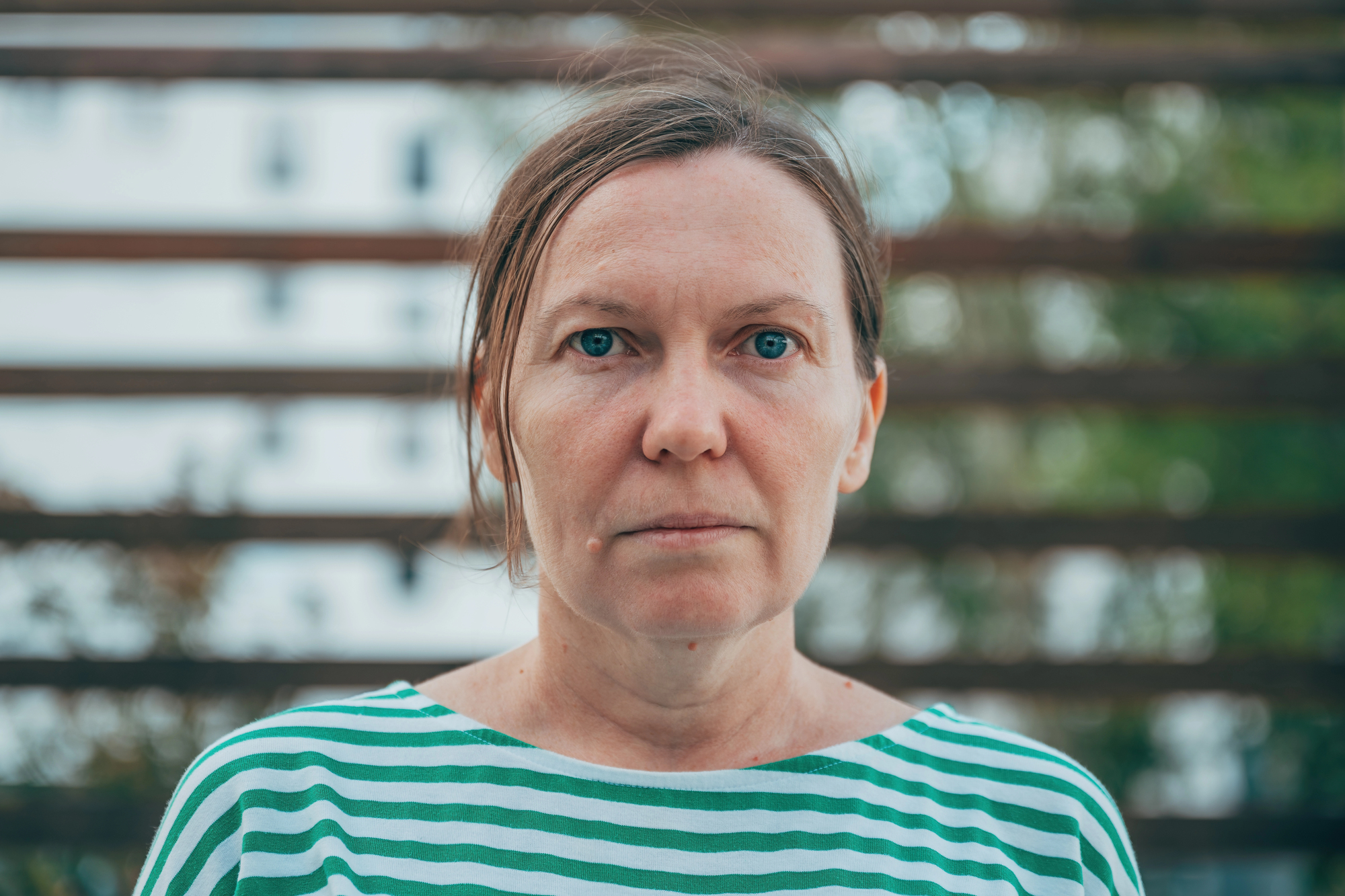 A woman with light skin and straight brown hair pulled back is wearing a green and white striped shirt. She stands outdoors in front of a blurred background of horizontal wooden slats and greenery, looking directly at the camera.