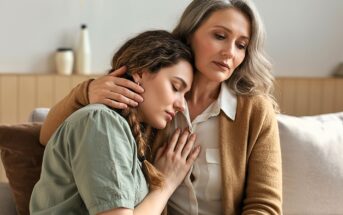 A woman with gray hair comforts a younger woman who rests her head on the older woman's chest. Both appear thoughtful and serious, sitting close together on a couch in a softly lit room.