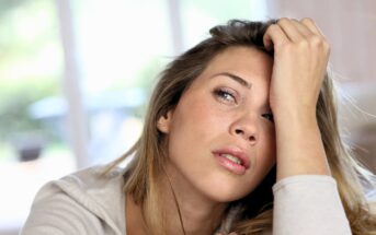A young woman with long blonde hair rests her head on her hand, looking tired or stressed. She is indoors with a blurred background, wearing a light-colored top.