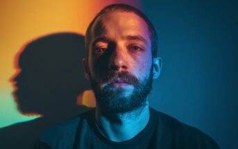 A man with a beard stands against a wall illuminated by dramatic yellow and blue lighting, casting a colorful shadow to his left and highlighting the contrast on his face.