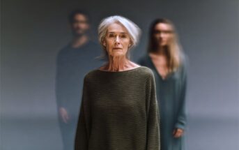 An elderly woman with gray hair stands in focus wearing a loose sweater, while a blurred man and woman stand behind her against a neutral background.