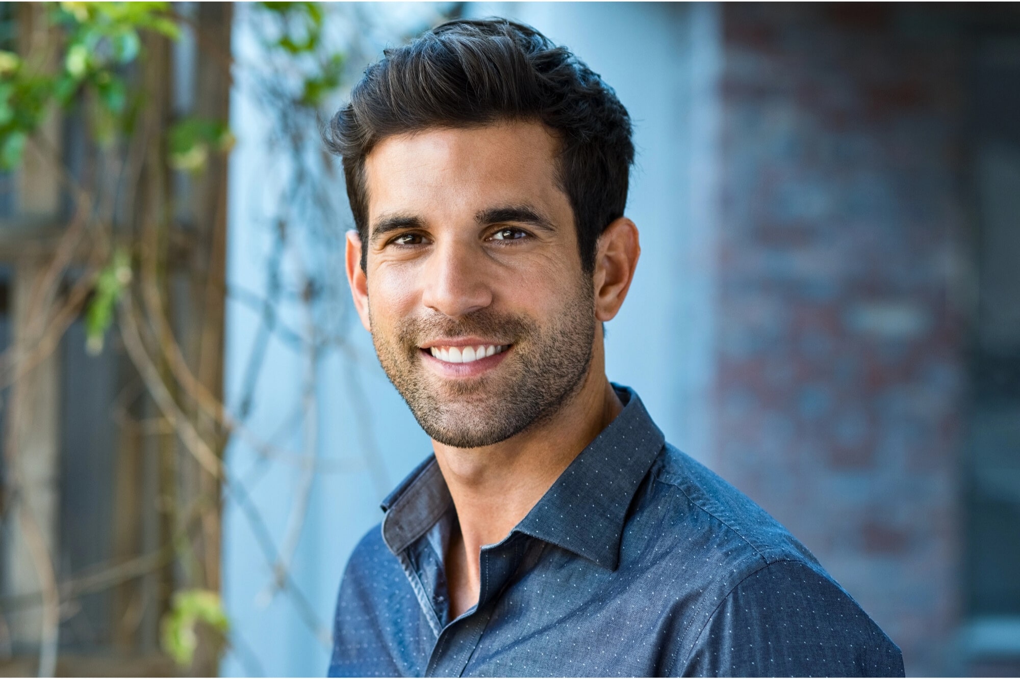 A man with short dark hair and a trimmed beard smiles at the camera. He is wearing a dark blue button-up shirt and standing outdoors next to a wall with greenery in the background.