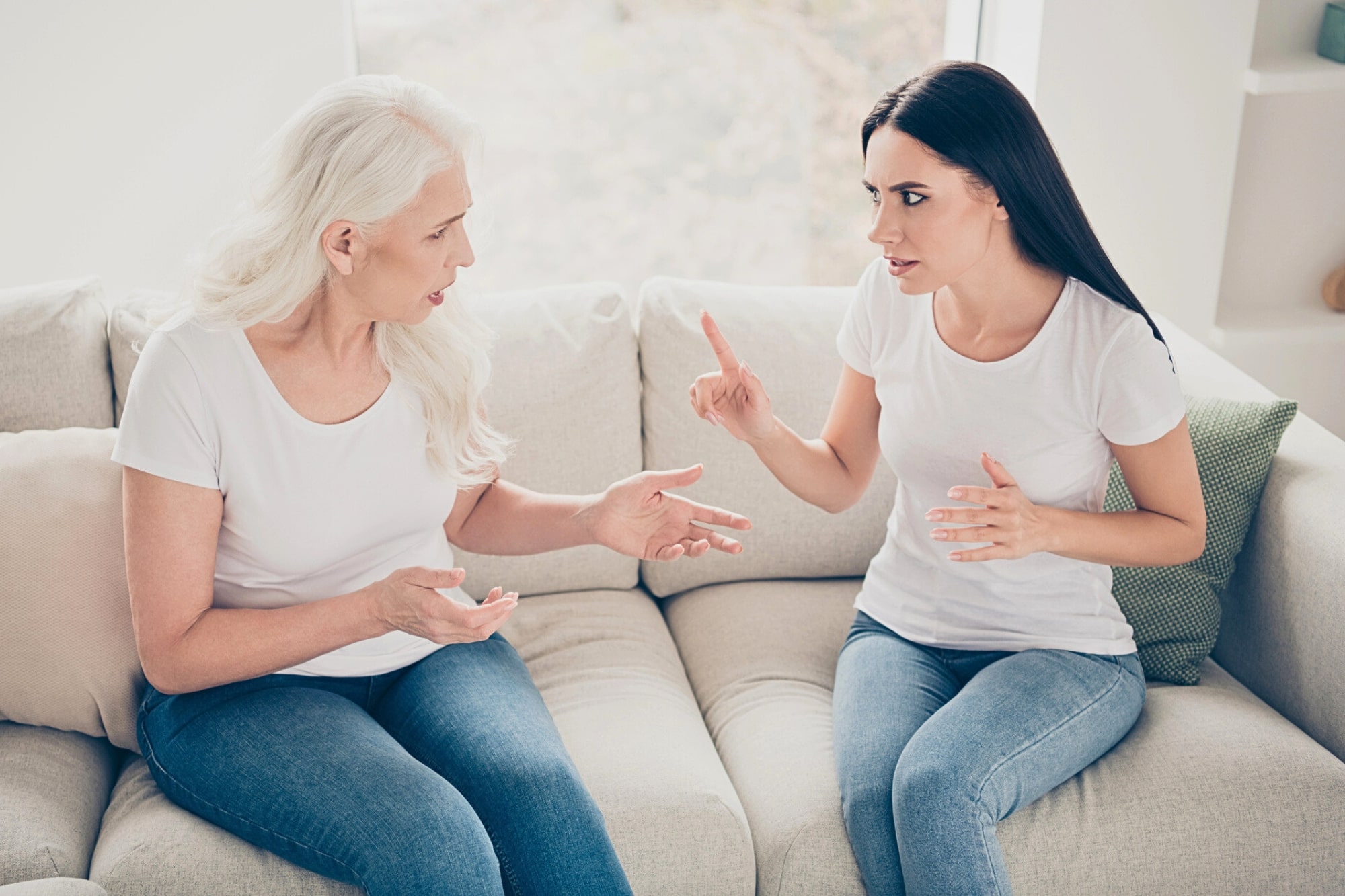 Two women sitting on a beige couch are having an intense conversation; the older woman gestures with open hands while the younger woman raises her index finger as if making a point. Both wear white tops and blue jeans.