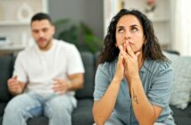 A woman sits on a couch looking frustrated and thoughtful, with her hands near her face, while a man in the background appears to be talking to her, looking concerned.