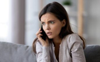 A young woman with long brown hair, wearing a light sweater, sits on a couch, looking worried while talking on a smartphone. Her brow is furrowed and her expression appears concerned or anxious.