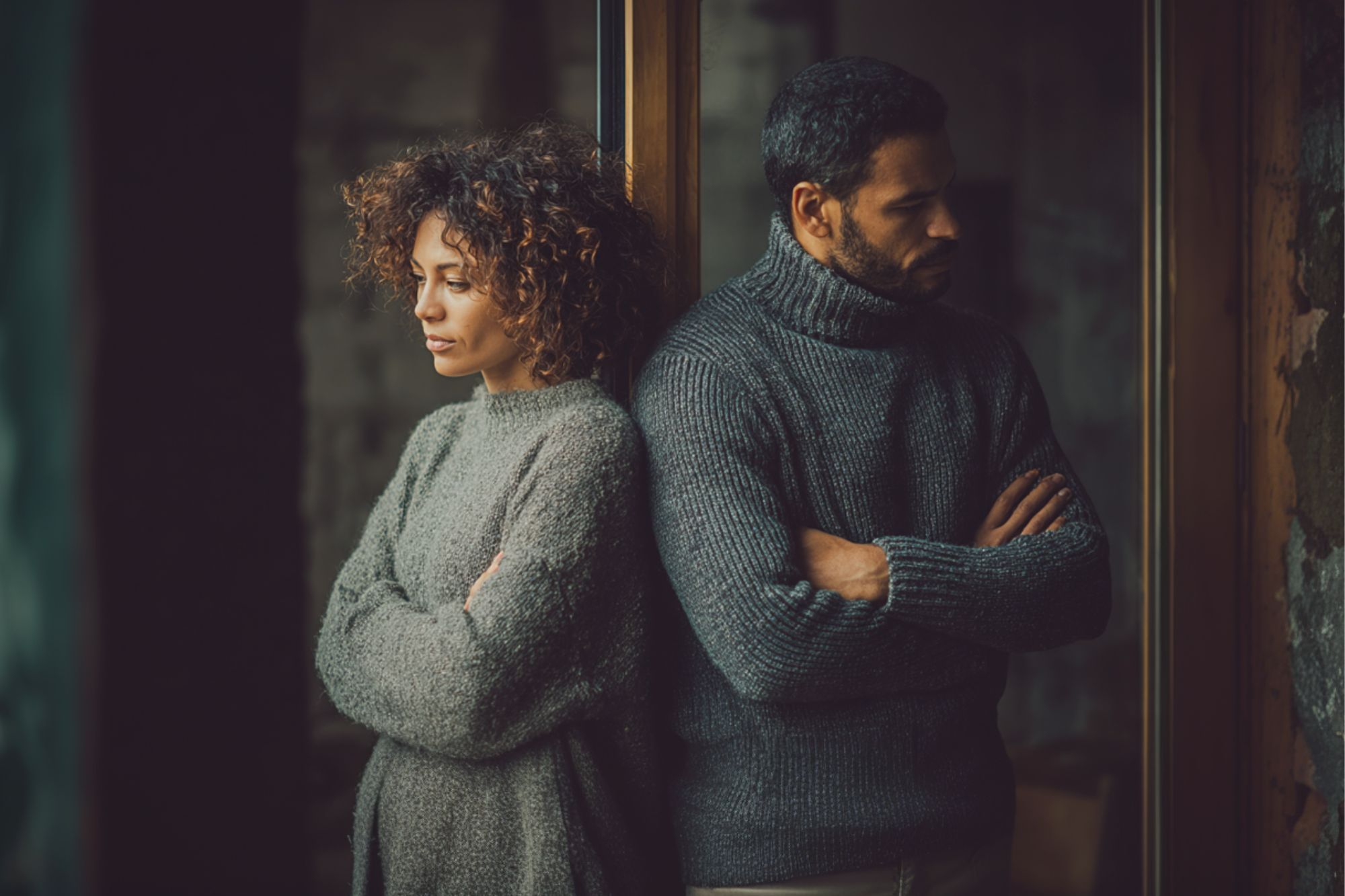 A woman and a man stand back to back with arms crossed, both looking away with serious expressions. They are indoors, wearing warm sweaters, and appear to be upset or in disagreement.