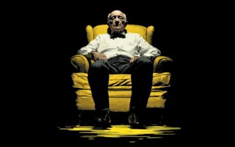 An elderly man in a white shirt and bow tie sits sternly in a yellow armchair, illuminated by dramatic lighting against a dark background.