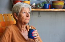 An older woman with short gray hair sips yerba mate from a blue cup using a metal straw while sitting on an orange chair. She wears a brown shirt and looks thoughtful, with potted plants visible in the background.