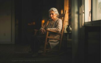 An elderly woman with gray hair sits thoughtfully in a wooden rocking chair by a window, soft light illuminating her face. She wears a beige sweater, and the room around her appears dim and calm.