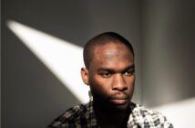 A man wearing a plaid shirt sits in soft sunlight, casting a triangle of light on the wall behind him. He looks down with a serious, contemplative expression, while shadows partially cover his face.