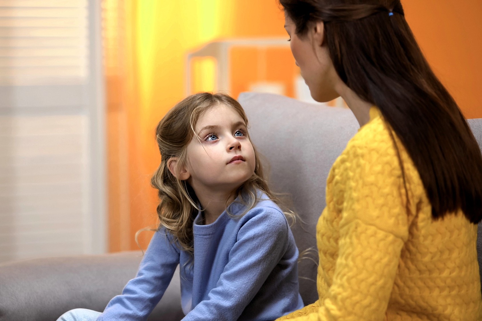 A young girl with blonde hair, wearing a light blue sweater, sits on a couch looking up attentively at a woman in a yellow sweater. The background is softly lit with warm yellow tones.