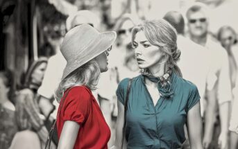 Two women face each other on a crowded street; one wears a red blouse and floppy hat, the other a teal blouse and scarf. The background is blurred, drawing focus to their intense expressions and interaction.