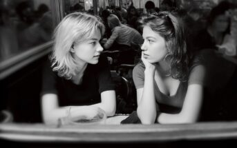 Two young women sit across from each other at a table in a busy café, gazing at each other and engaged in conversation. The scene is in black and white, with blurred people in the background.