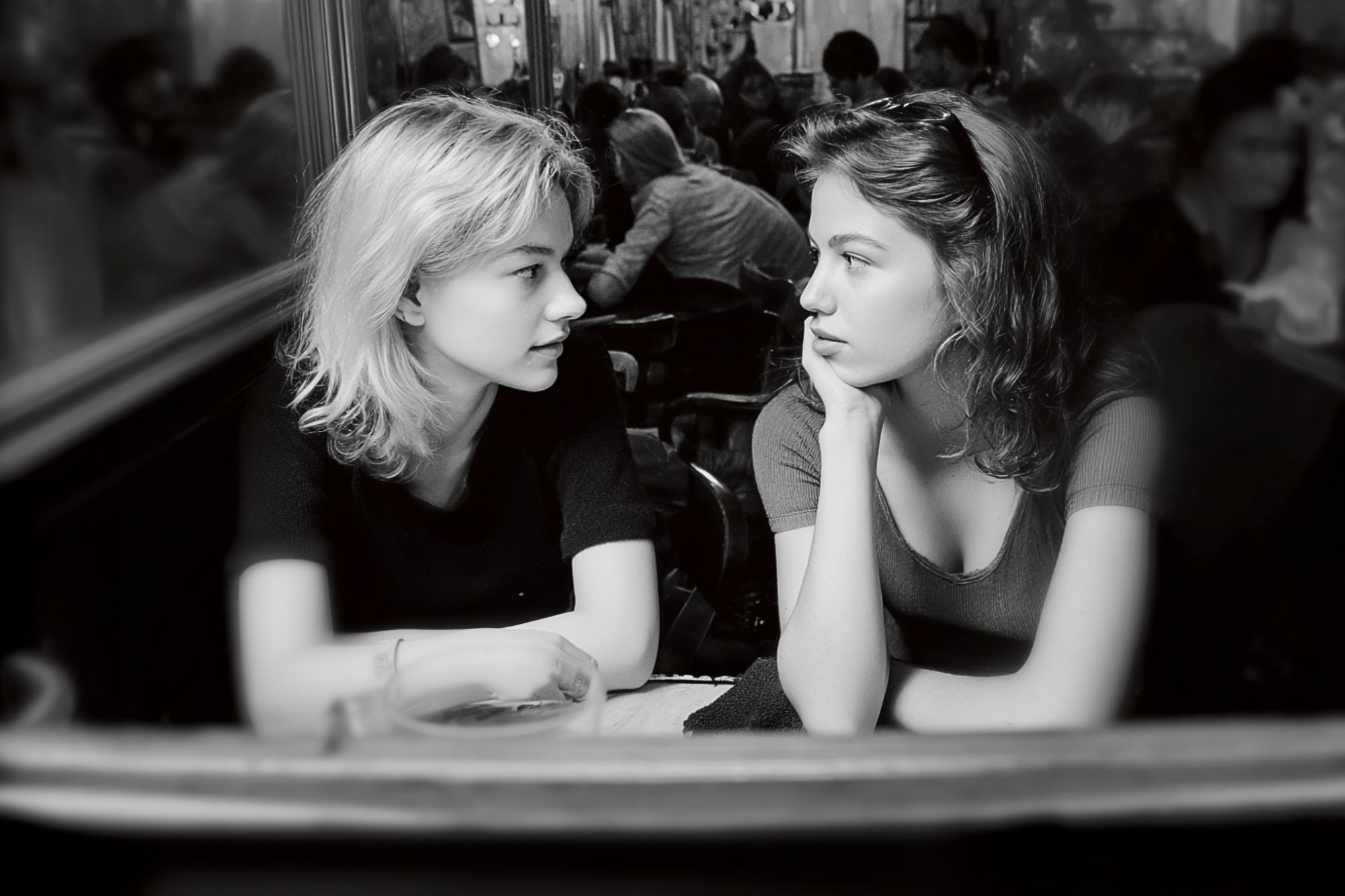 Two young women sit across from each other at a table in a busy café, gazing at each other and engaged in conversation. The scene is in black and white, with blurred people in the background.