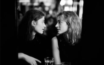Two women sit closely together at a table in a dimly lit setting, engaging in an intimate conversation. Glasses of water are on the table in front of them.