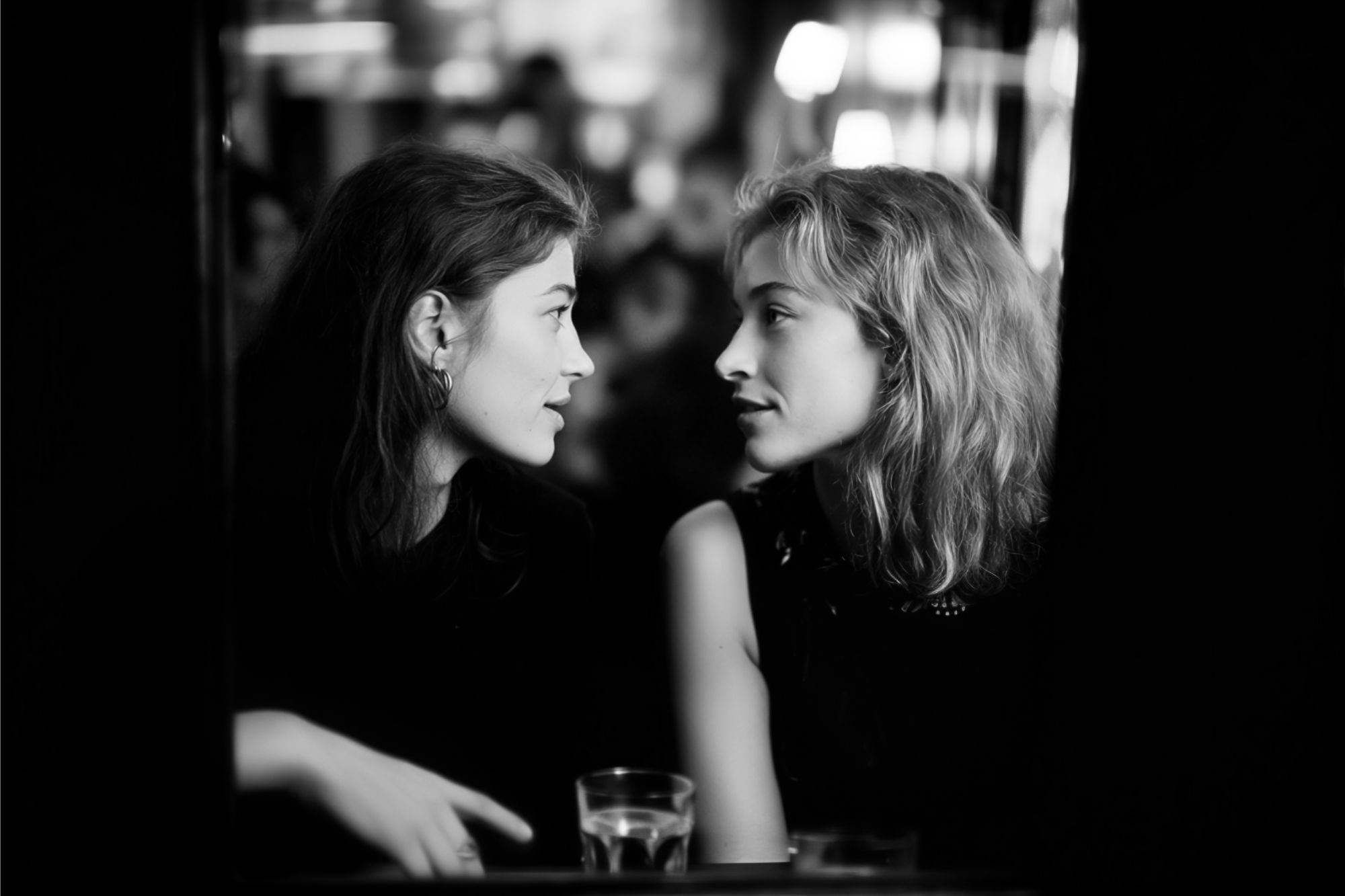 Two women sit closely together at a table in a dimly lit setting, engaging in an intimate conversation. Glasses of water are on the table in front of them.