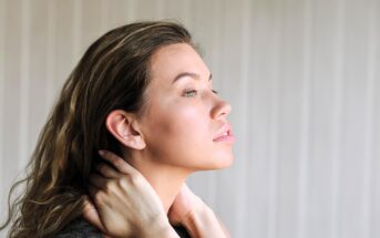 A woman with long brown hair gazes to the side, holding her hands gently around her neck. She has a calm expression and stands in front of a light-colored, vertically-striped background.