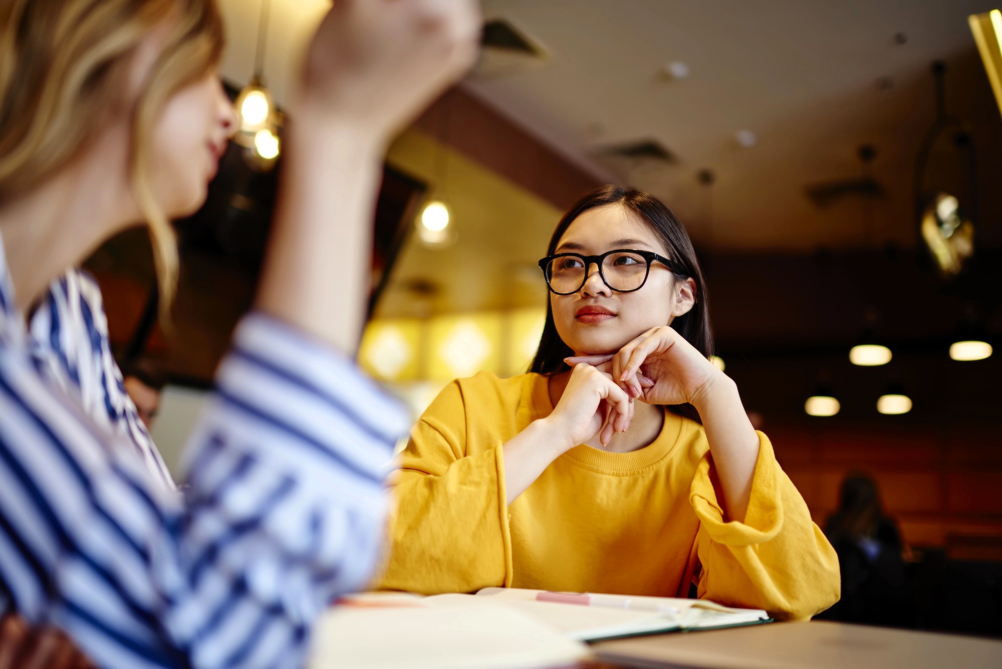 Two young women sit at a table indoors, talking. One woman wearing glasses and a yellow top listens attentively with her hands under her chin, while the other gestures with her hand. Books and papers are on the table.