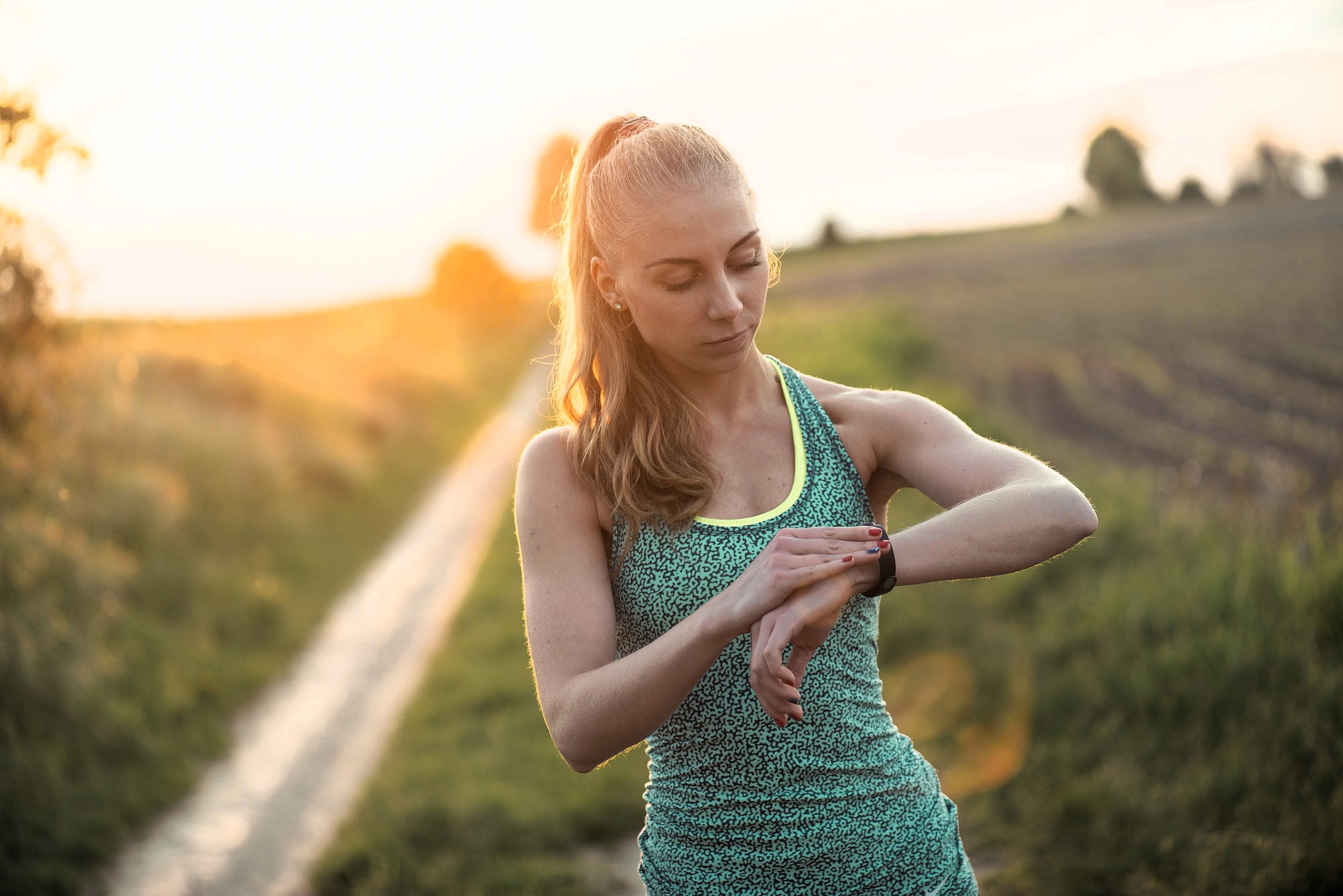 A woman in athletic wear stands on a rural dirt path at sunset, looking down and checking her smartwatch, with fields and soft sunlight in the background.