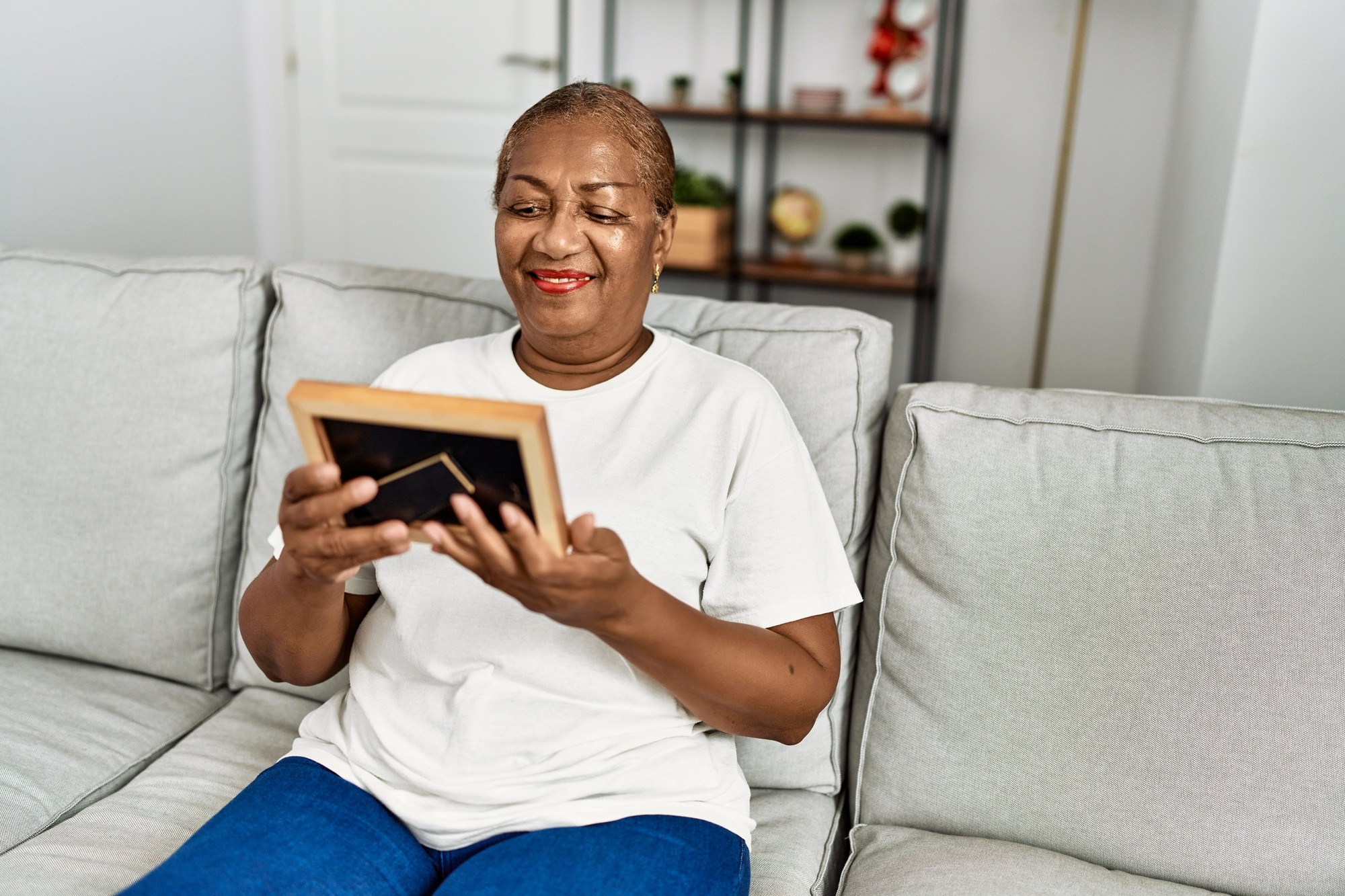 An older woman sits on a light-colored sofa, smiling warmly as she looks at a framed photo she holds in her hands. Shelving with plants and decor is visible in the softly lit background.
