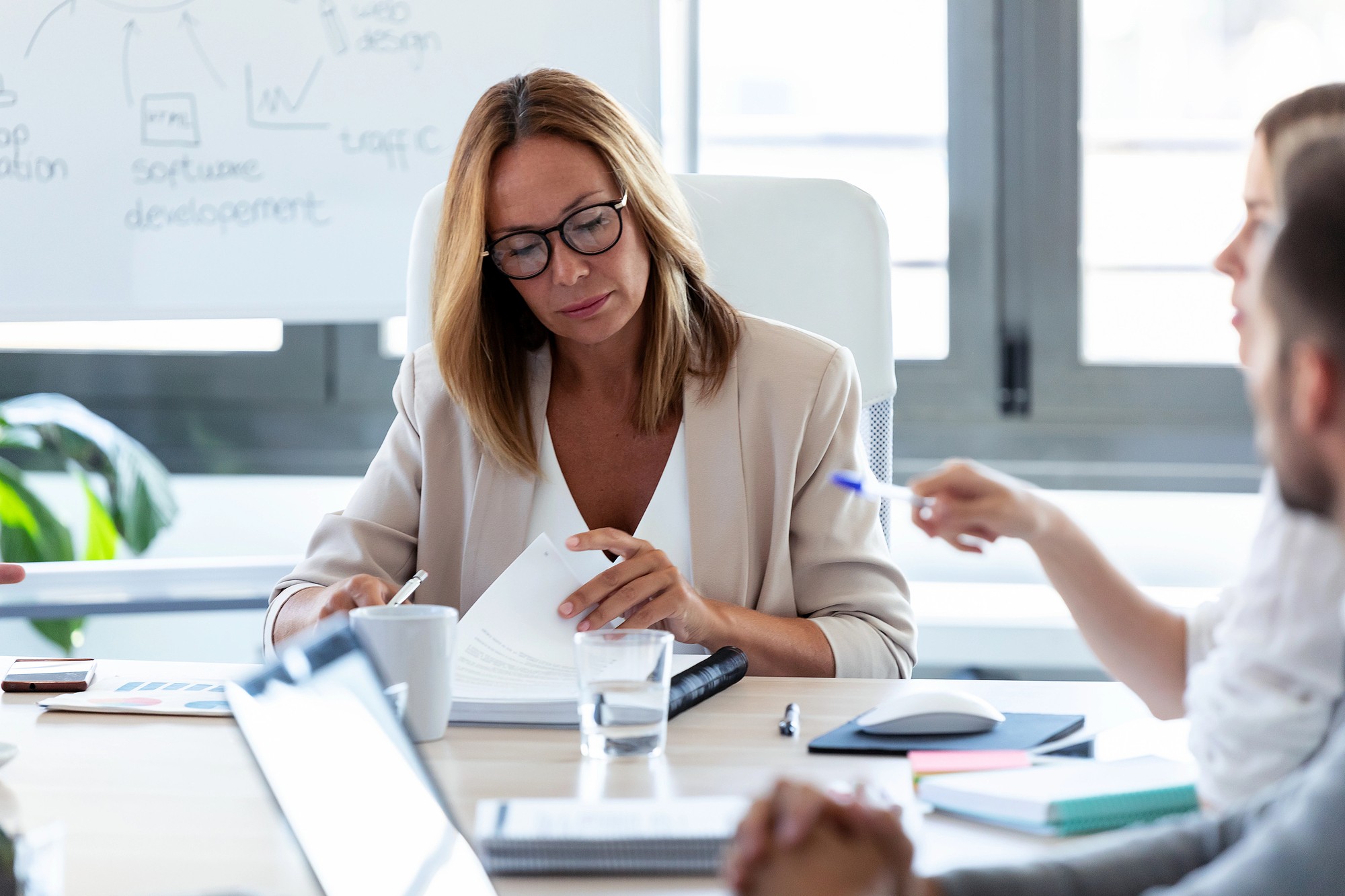 A woman wearing glasses sits at a desk in an office, reading documents. Two colleagues are partially visible, engaging in discussion. There are papers, a laptop, a glass of water, and a whiteboard with diagrams in the background.
