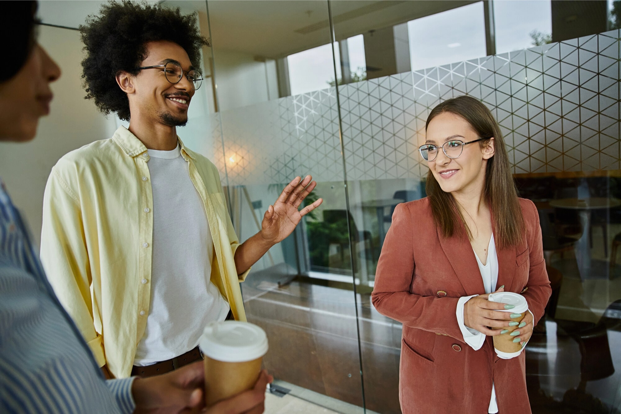 Three people stand indoors holding takeaway coffee cups, engaged in conversation. One man gestures while smiling, and a woman in a rust blazer listens and smiles. They are near a glass wall with a geometric pattern.