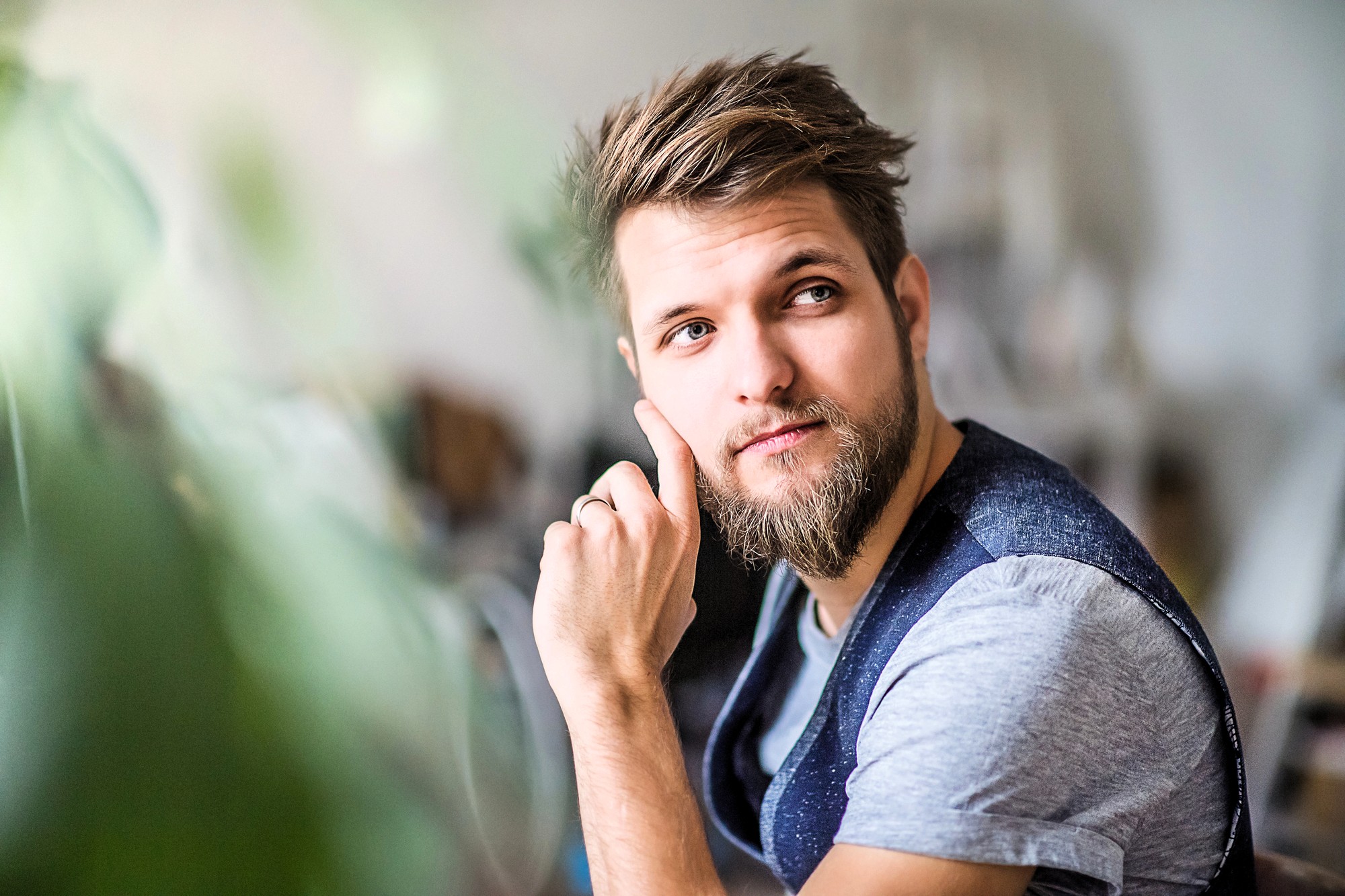 A young man with a beard and mustache, wearing a gray t-shirt and dark vest, sits indoors with his hand resting on his cheek, looking thoughtfully to the side. Green foliage is blurred in the foreground.