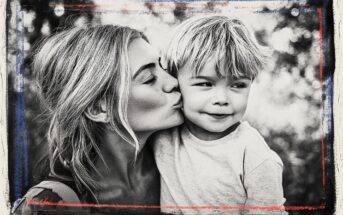 A woman kisses a young boy on the cheek outdoors. Both have light hair, and the boy smiles softly. The black-and-white image is framed with a rough border featuring blue and red lines.