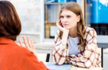 A young woman in a plaid shirt listens attentively with her chin resting on her hand as another person, seen from behind, gestures during an outdoor conversation at a table.