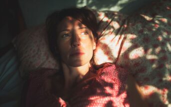 A woman lies on a floral pillow, looking thoughtfully into the distance. Sunlight and shadows from leaves create patterns on her face and red patterned top.
