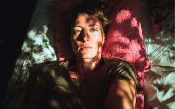 A person with short hair lies on a bed, head on a pink pillow, looking up with a serious expression. Sunlight creates dramatic shadows across their face and upper body.