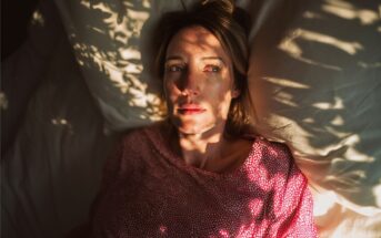 A woman lies on a bed with white pillows, wearing a pink top. Sunlight filters through leaves, casting shadows on her face. She appears thoughtful and looks off to the side.