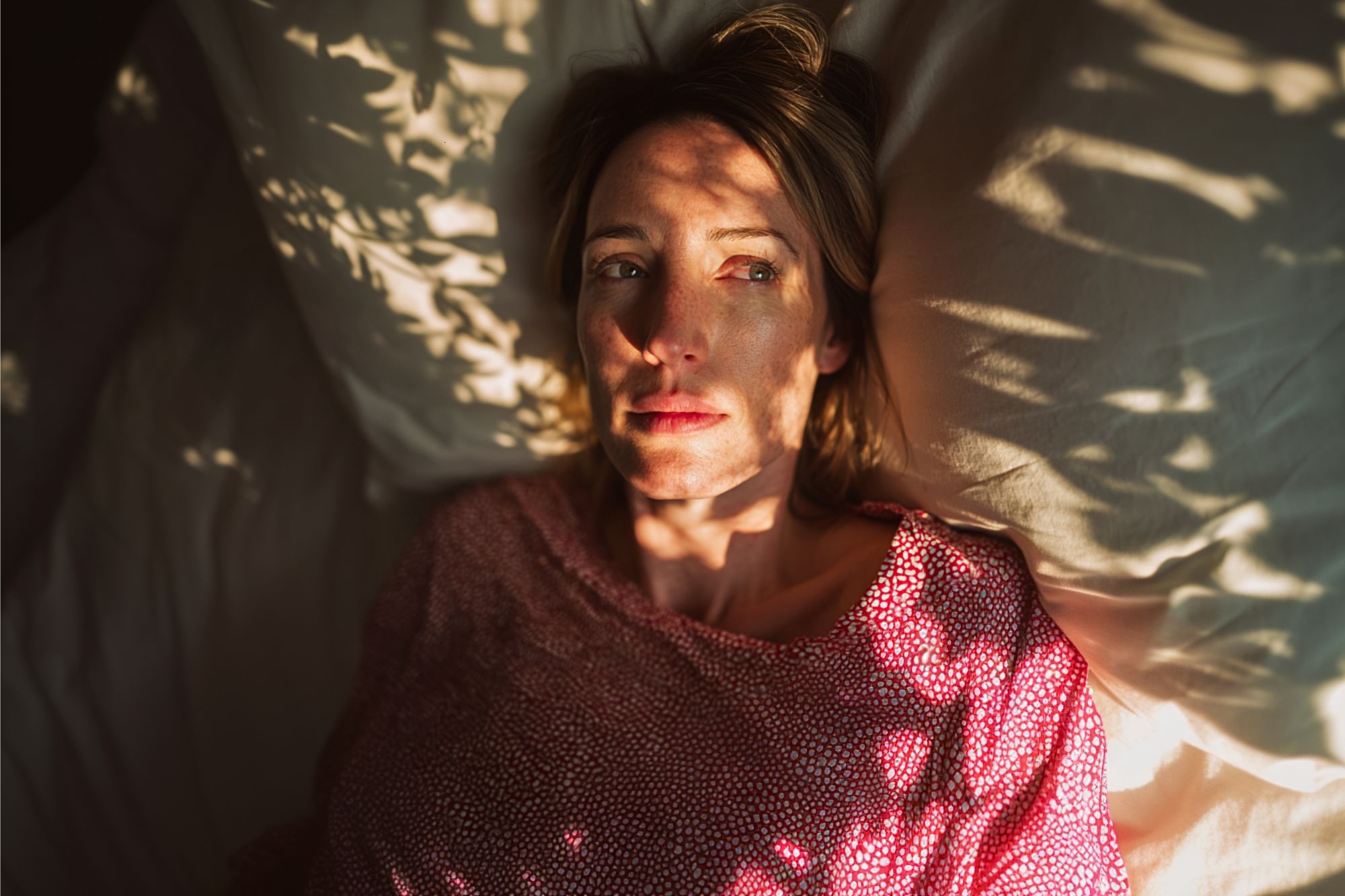 A woman lies on a bed with white pillows, wearing a pink top. Sunlight filters through leaves, casting shadows on her face. She appears thoughtful and looks off to the side.