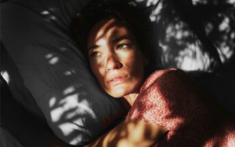 A woman with freckles lying on a pillow, with dappled light casting shadows across her face.