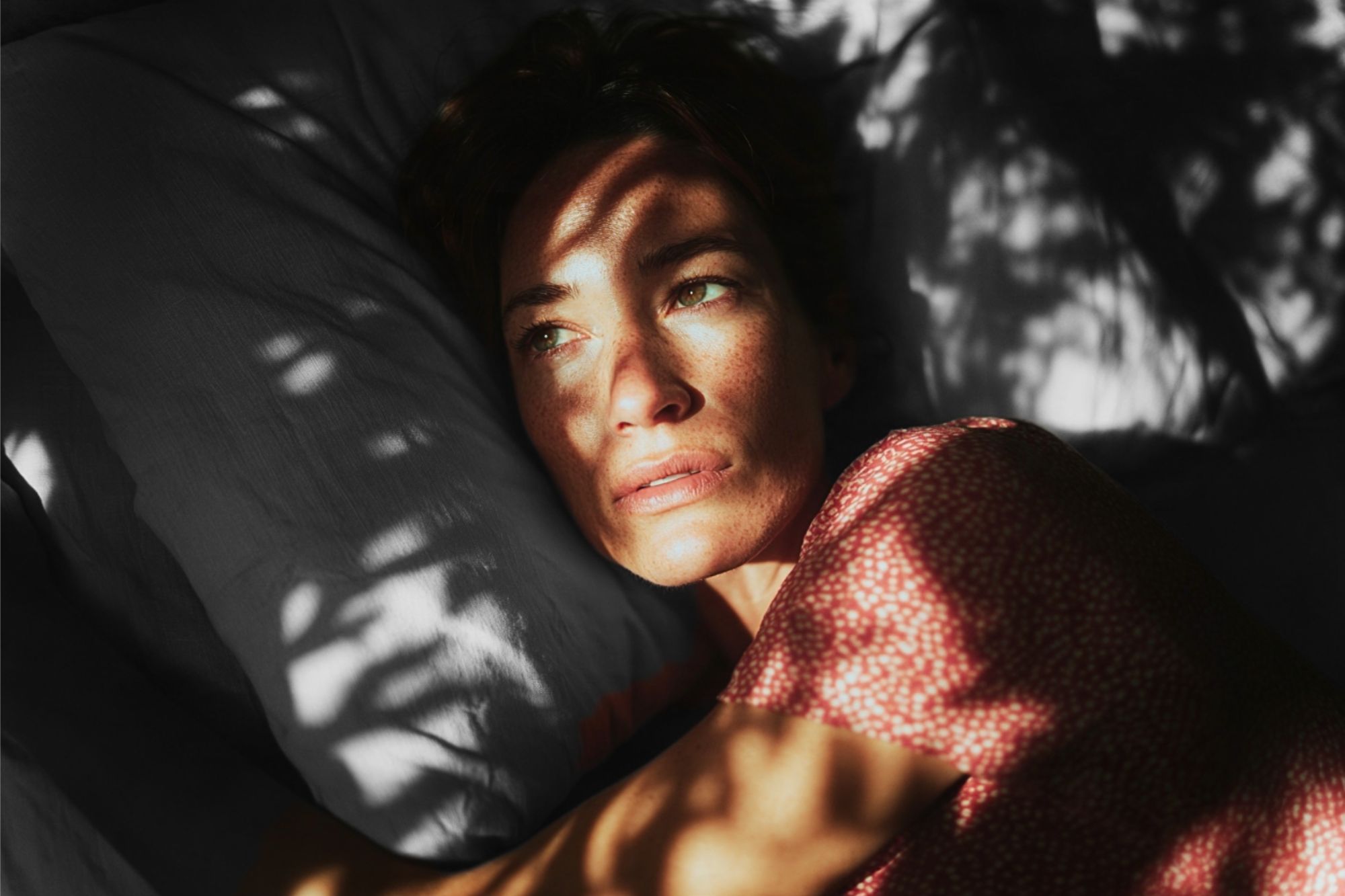 A woman with freckles lying on a pillow, with dappled light casting shadows across her face.