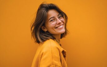 A young woman with shoulder-length brown hair, wearing round glasses and a yellow jacket, smiles while standing in front of a solid orange background.