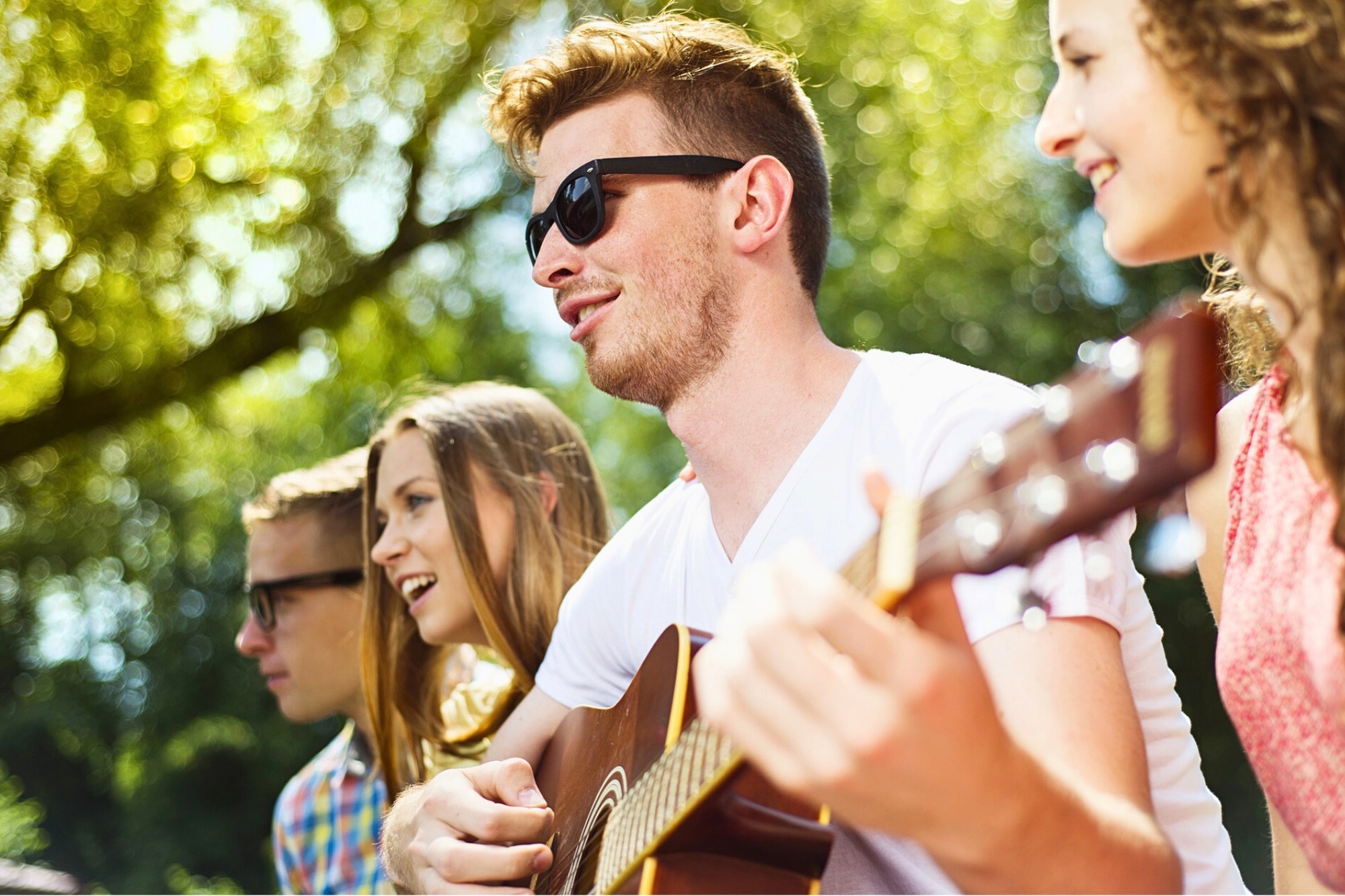 Four young adults sit outdoors on a sunny day. One person plays an acoustic guitar while the others smile and sing along. All are wearing casual clothes, and two of them wear sunglasses. Trees and greenery are in the background.